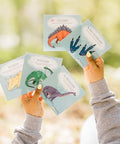 Des mains montrant des cartes d'apprentissage actives dinosaures (tricératops, stégosaure, brontosaure)