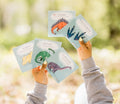 Des mains montrant des cartes d'apprentissage actives dinosaures (tricératops, stégosaure, brontosaure)